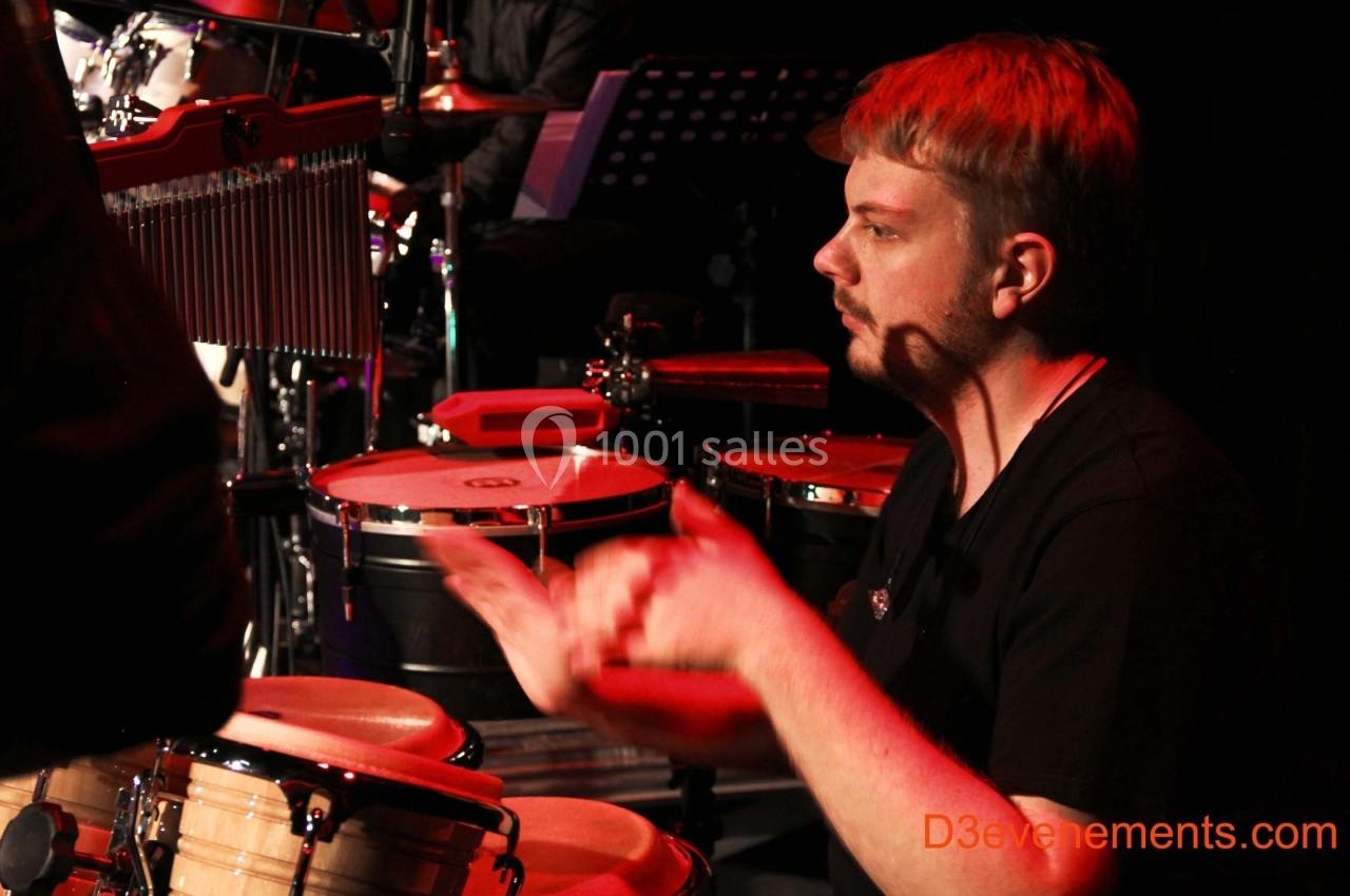 Un musicien joue des percussions sur scène, éclairé par une lumière rouge.