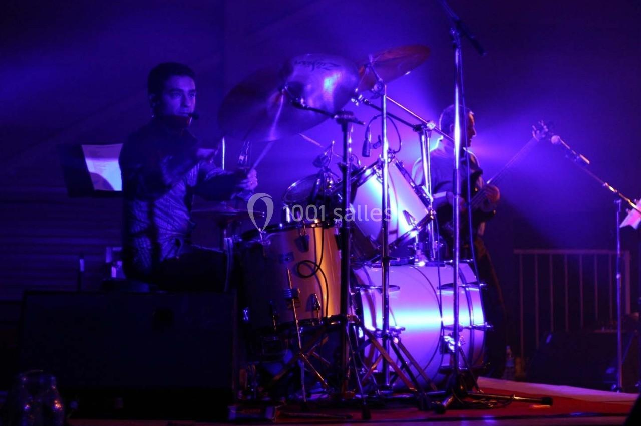 Un batteur joue sur une batterie éclairée par des lumières violettes lors d'un concert en intérieur.