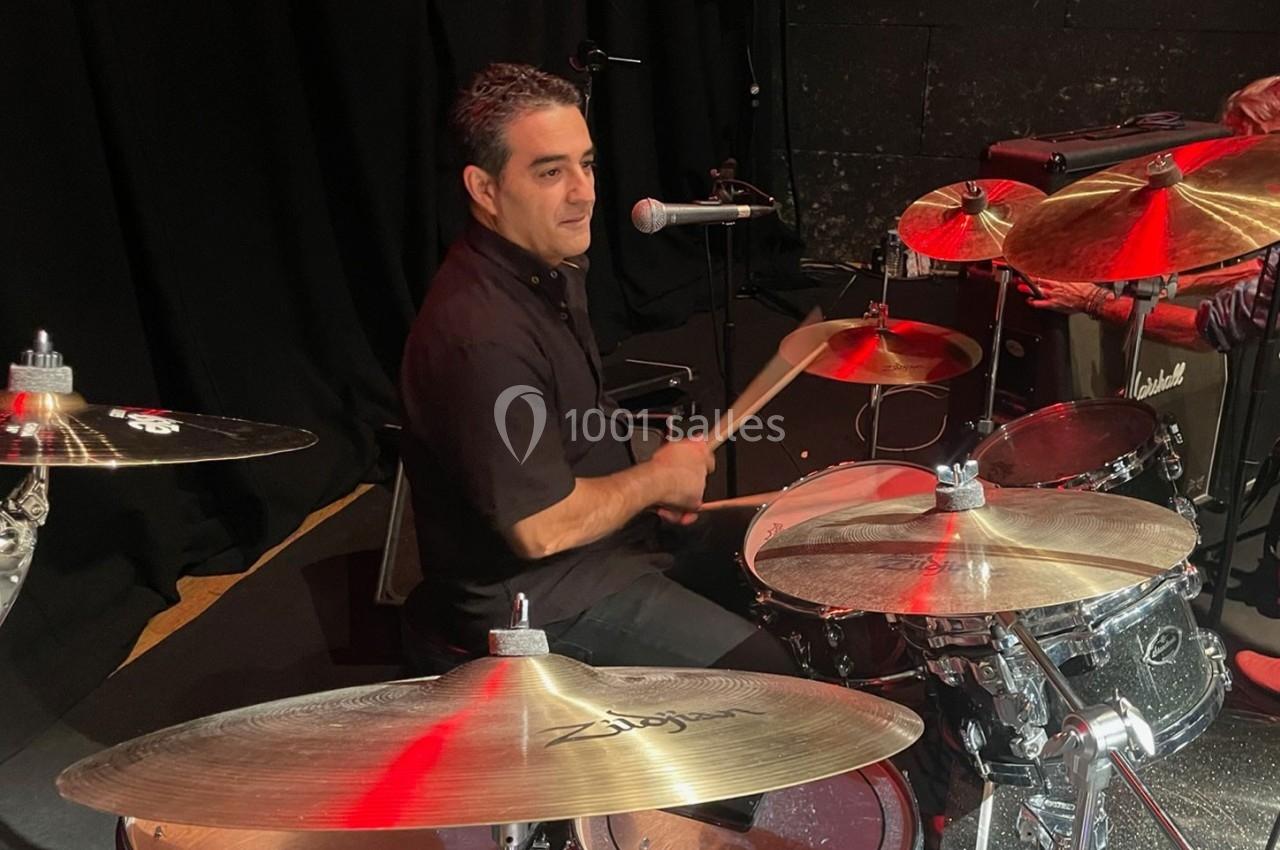 Un batteur joue sur une batterie argentée sur une scène sombre, avec un micro à proximité.