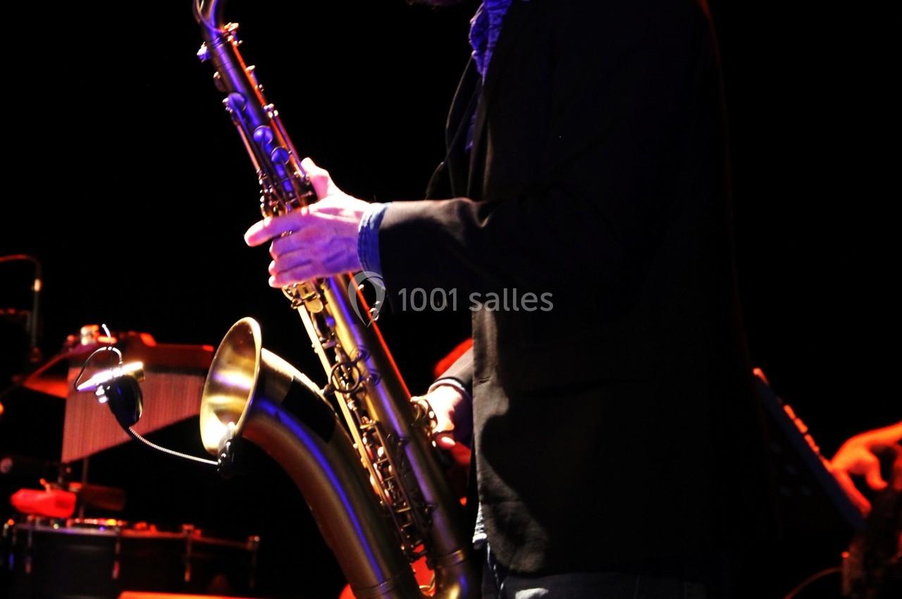 Un musicien joue du saxophone sur scène, avec des percussions en arrière-plan dans une ambiance sombre.
