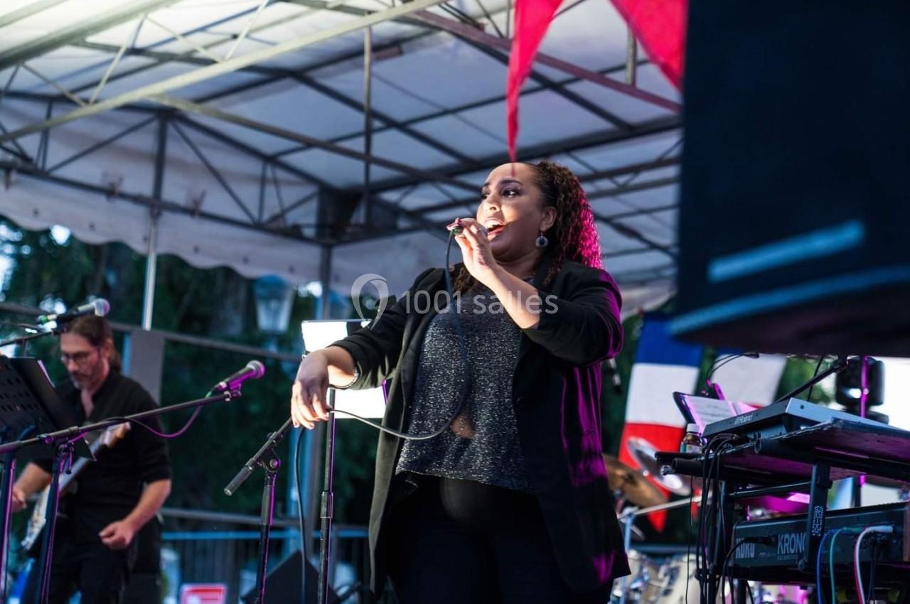 Une chanteuse sur scène tenant un micro, accompagnée de musiciens, sous une structure couverte en extérieur.
