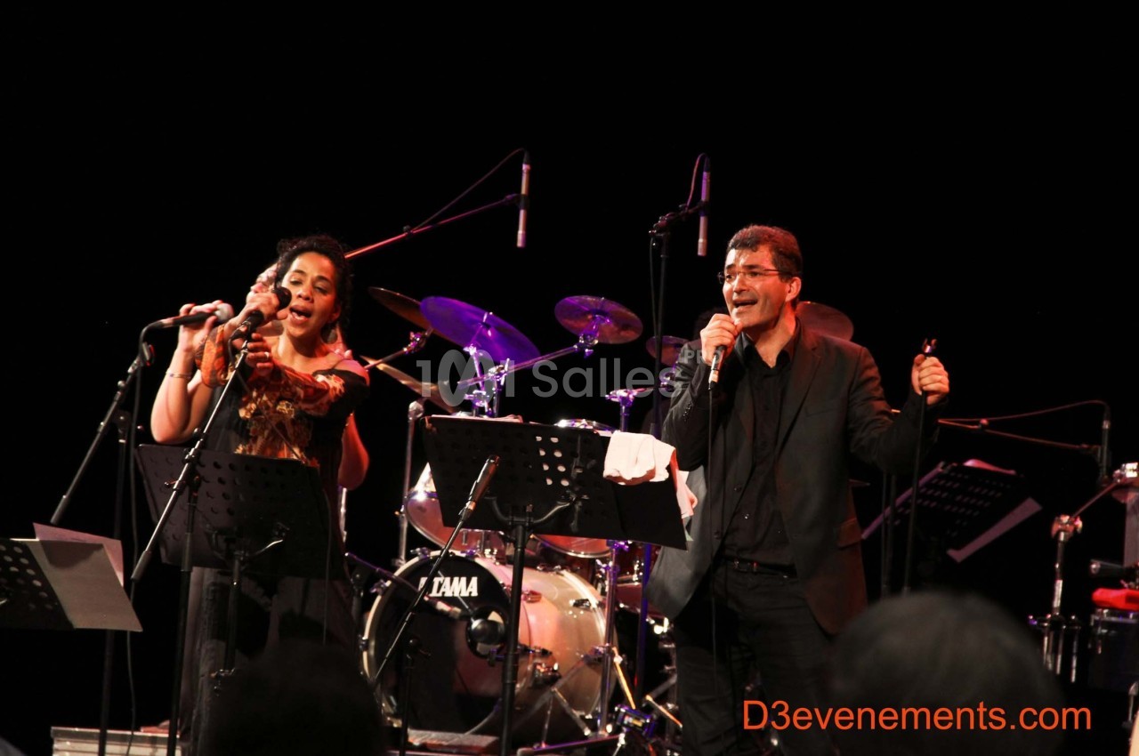 Deux chanteurs sur scène interprétant une chanson, accompagnés de musiciens et d'instruments en arrière-plan.