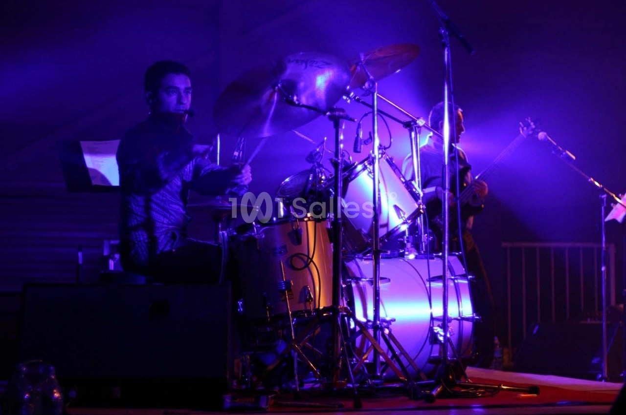 Un batteur joue sur une batterie éclairée par des lumières violettes lors d'un concert en intérieur.