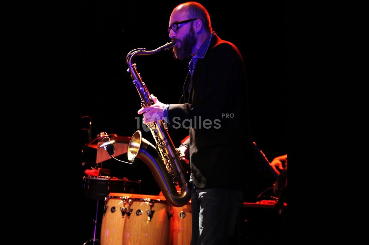 Un musicien joue du saxophone sur scène, éclairé par des lumières, avec des percussions en arrière-plan.