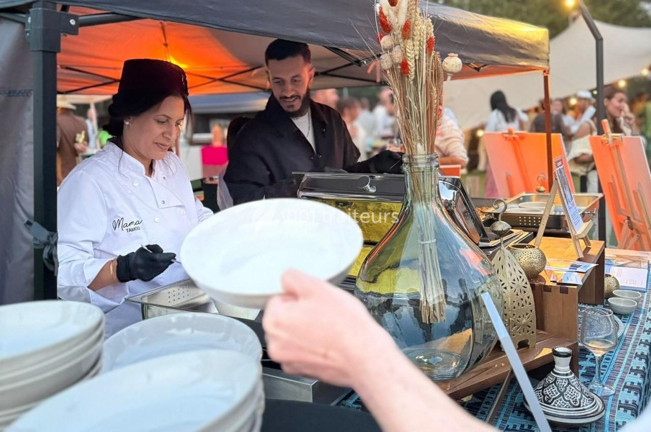 Une cheffe prépare des plats sous un stand de marché, avec un homme à ses côtés et des clients en arrière-plan.