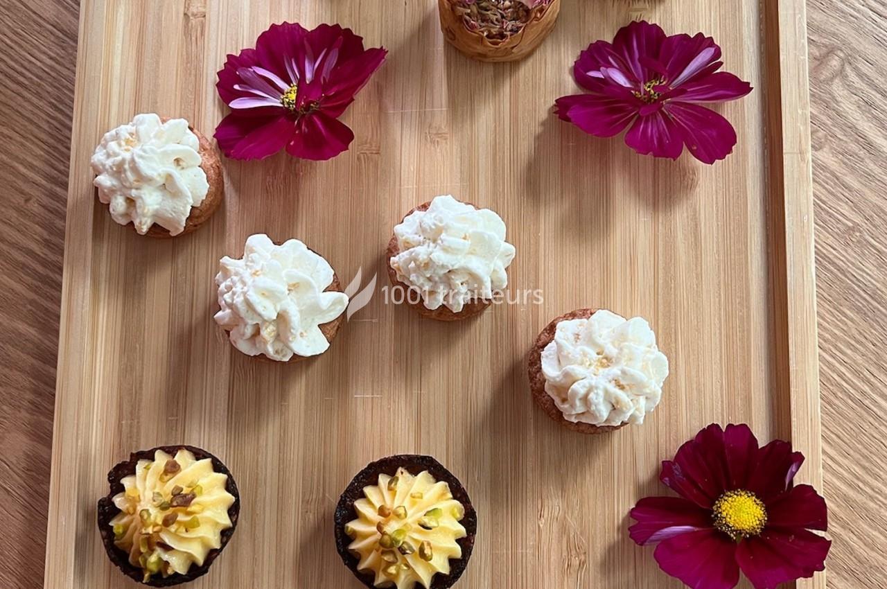 Plateau en bois avec des petits gâteaux décorés de crème et des fleurs roses disposées autour.