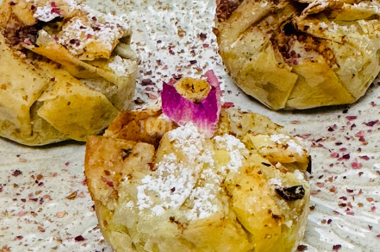 Feuilletés dorés garnis, décorés de sucre glace et de pétales de rose, posés sur une assiette parsemée d'épices.