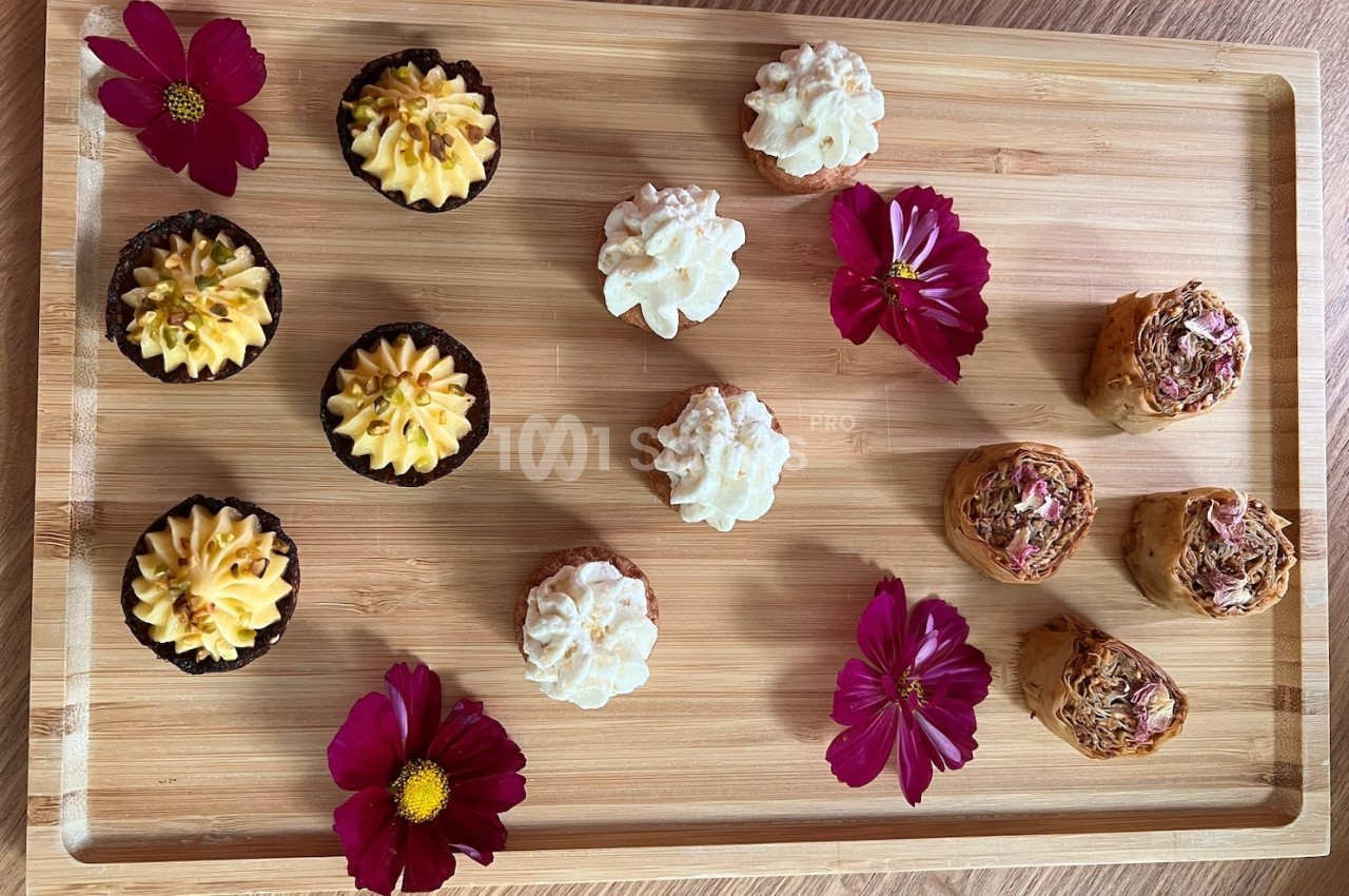 Plateau en bois avec des petits gâteaux variés décorés de crème et de fleurs comestibles, entourés de fleurs roses.