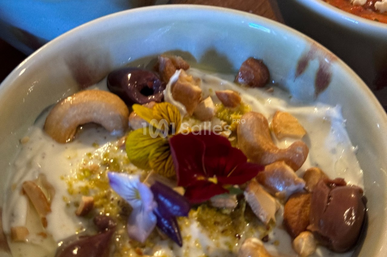 Bol de yaourt garni de noix de cajou, olives, fleurs comestibles et zestes, présenté dans une vaisselle en céramique.