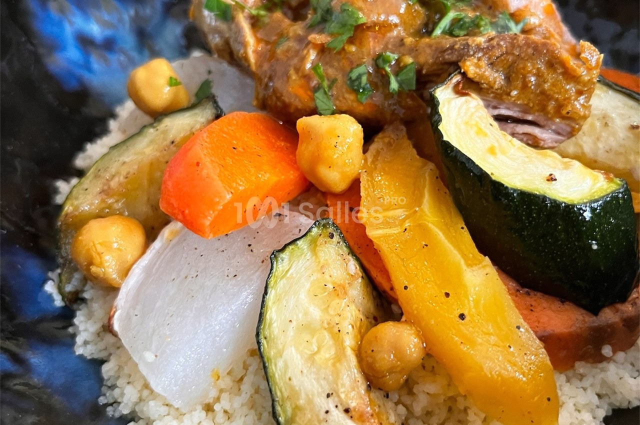 Plat de couscous garni de légumes colorés, pois chiches et viande en sauce, servi dans une assiette noire.