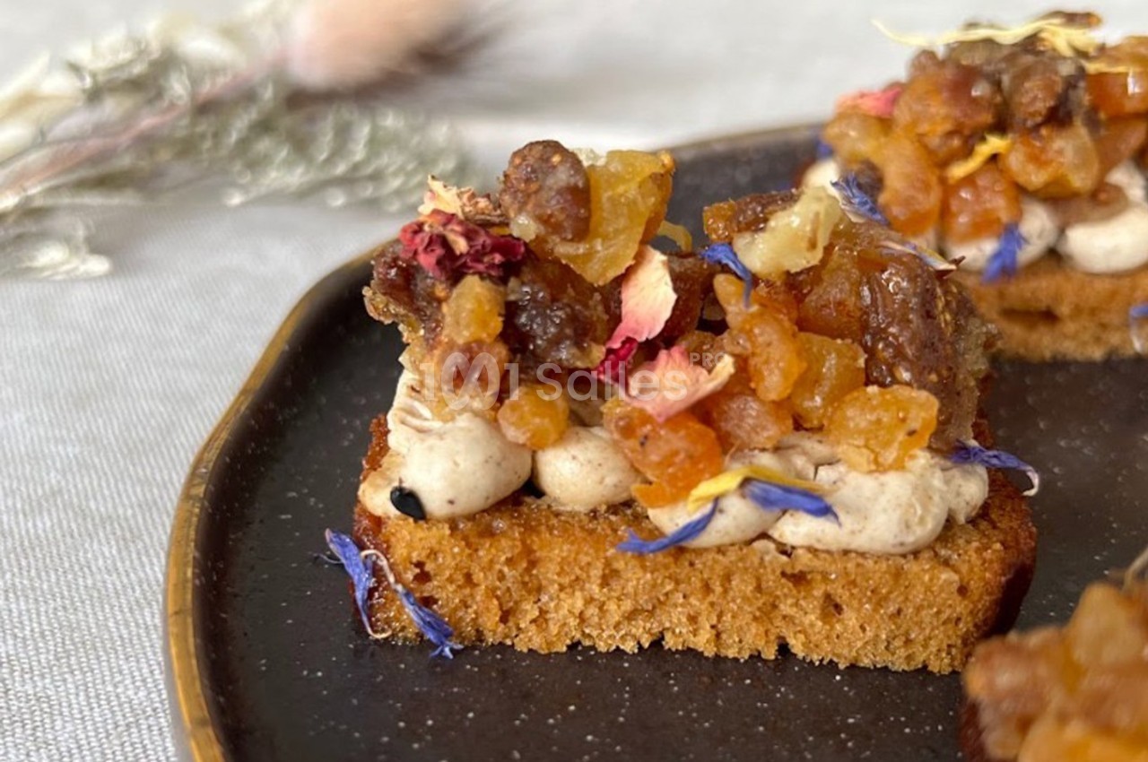 Tranche de pain garnie de crème et de fruits secs, décorée de fleurs comestibles, sur une assiette sombre.