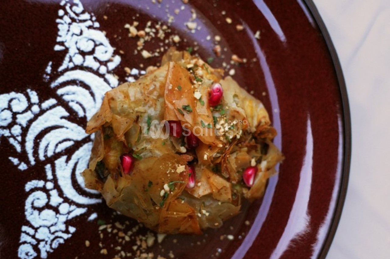 Feuilleté doré garni de graines de grenade, herbes fraîches et éclats de noix, présenté sur une assiette décorative.