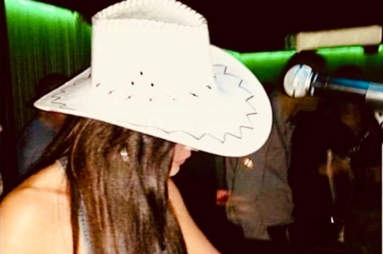 Femme portant un chapeau blanc de style cowboy, penchée près d'un microphone dans un environnement sombre.