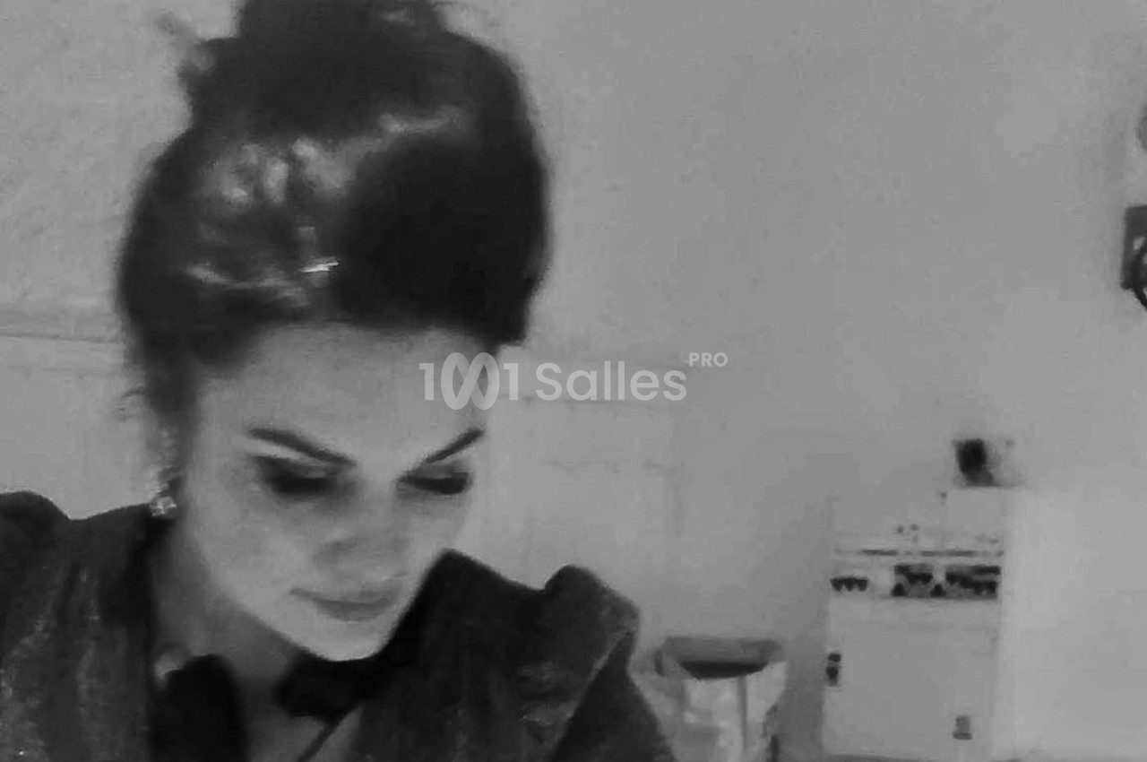 Femme aux cheveux relevés, regard baissé, dans une pièce sobrement éclairée avec des meubles en arrière-plan.