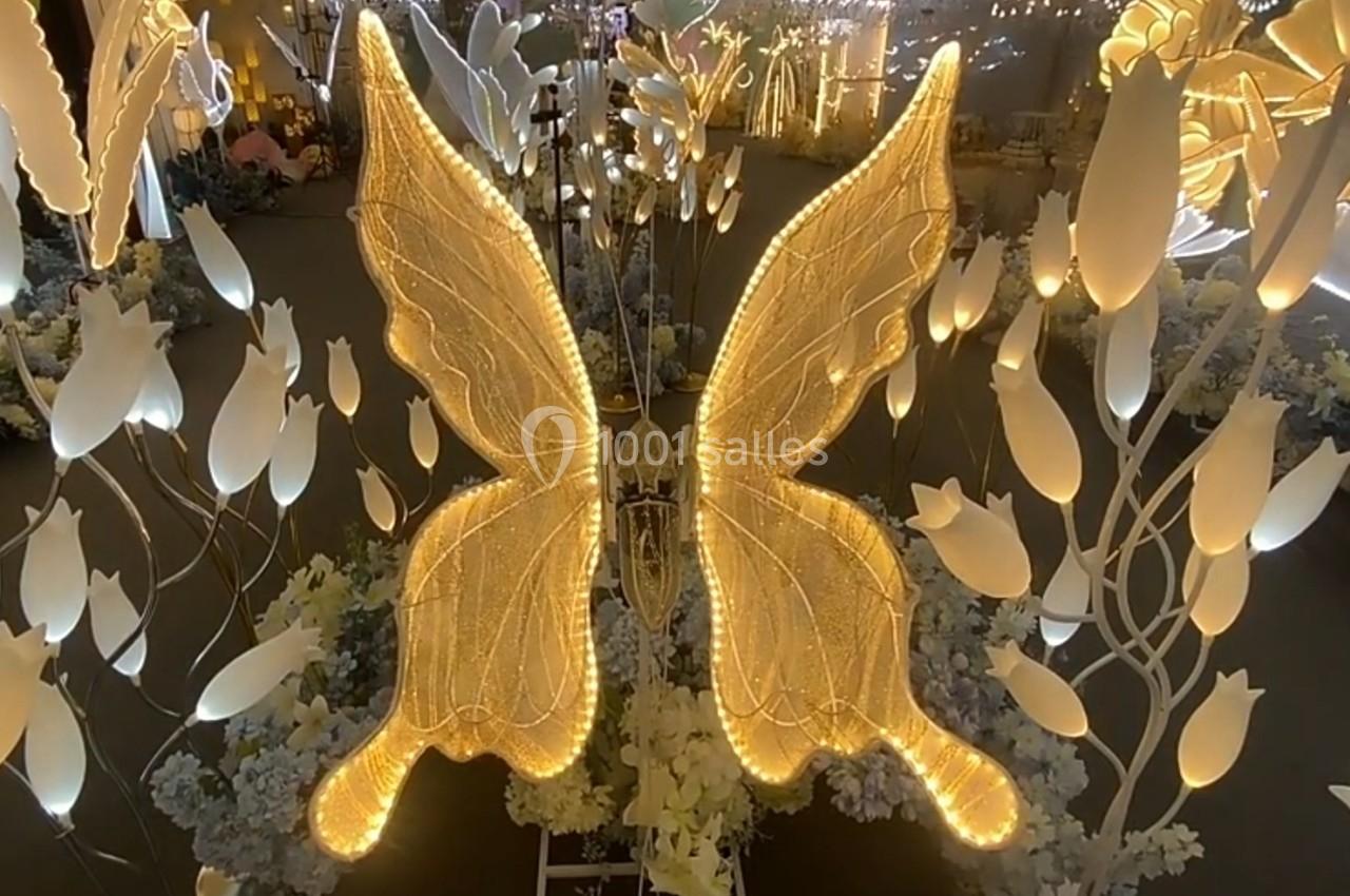 Papillon lumineux doré entouré de fleurs blanches illuminées dans un décor nocturne.