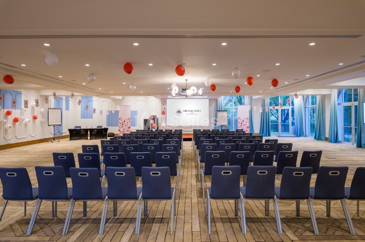 Salle de conférence avec des rangées de chaises, un écran de projection, des décorations rouges et blanches.