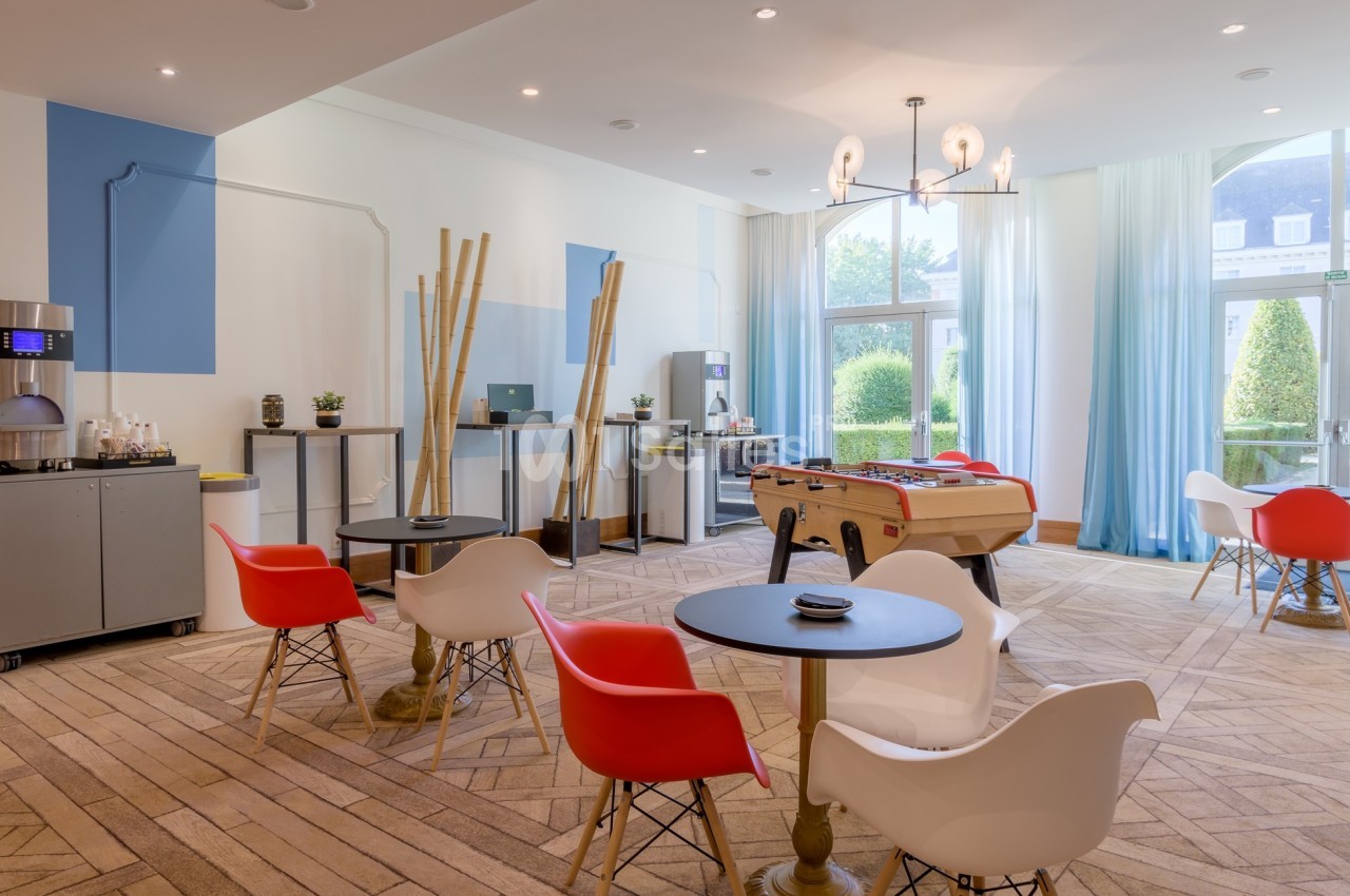 Salle lumineuse avec tables, chaises colorées, baby-foot, machine à café et grandes fenêtres donnant sur un jardin.