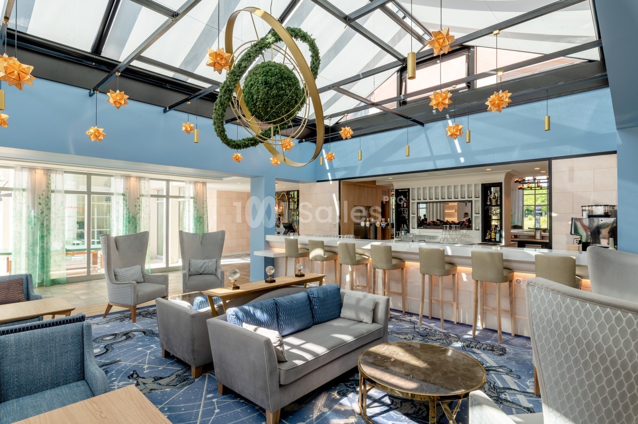 Espace lounge lumineux avec verrière, mobilier moderne, bar en arrière-plan et décoration suspendue végétale.