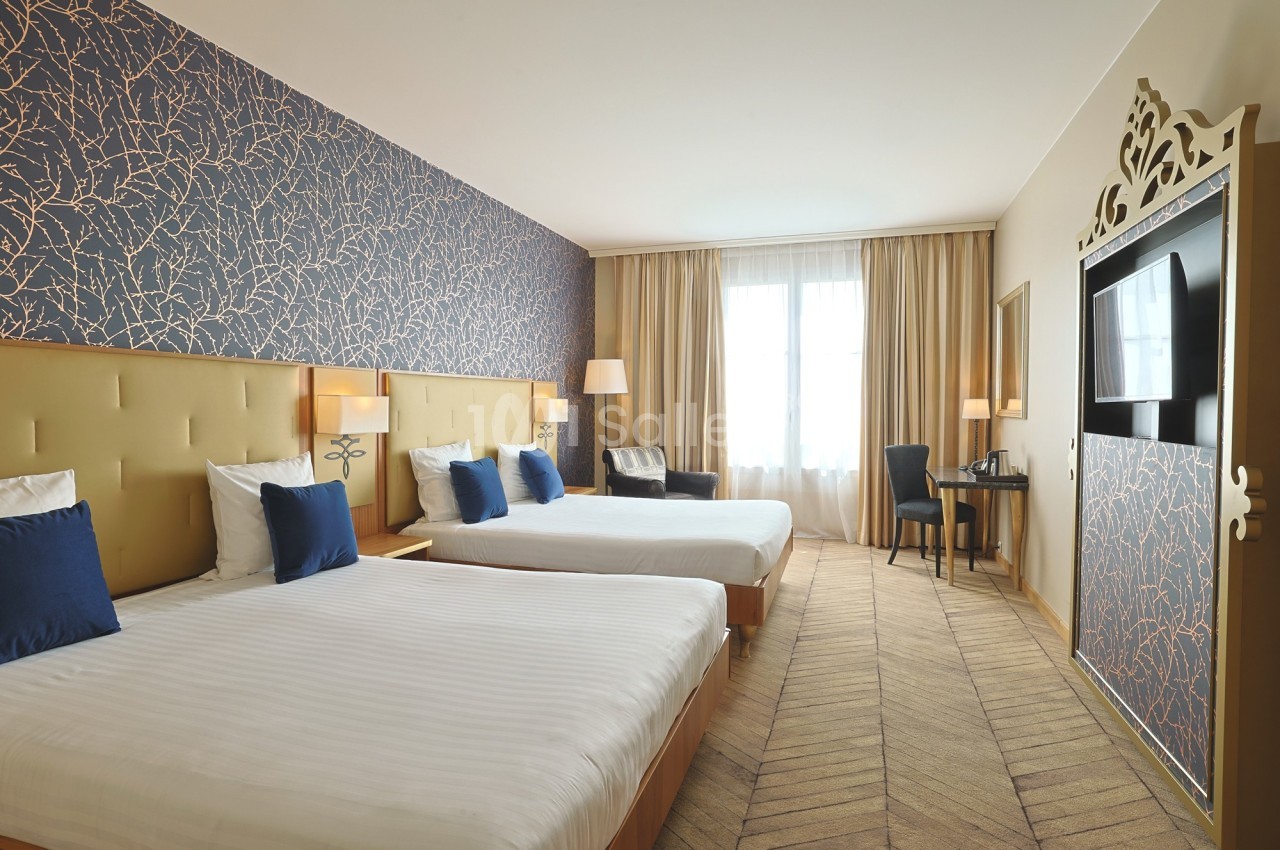 Chambre d'hôtel lumineuse avec deux lits doubles, décor moderne, bureau, télévision et grande fenêtre avec rideaux.