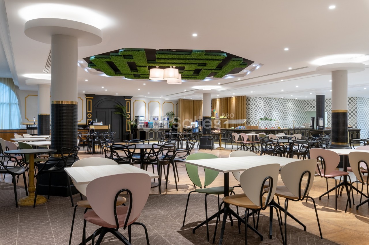 Salle de restaurant moderne avec tables et chaises colorées, plafond décoratif et buffet en arrière-plan.