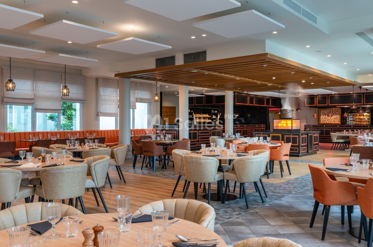 Salle de restaurant moderne avec tables dressées, chaises rembourrées et cuisine ouverte en arrière-plan.