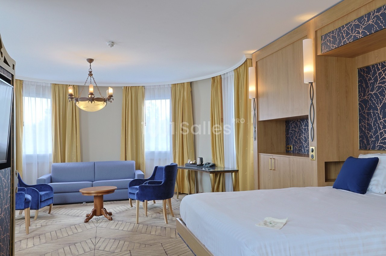 Chambre d'hôtel lumineuse avec lit double, coin salon, rideaux jaunes, mobilier en bois et fauteuils bleus.
