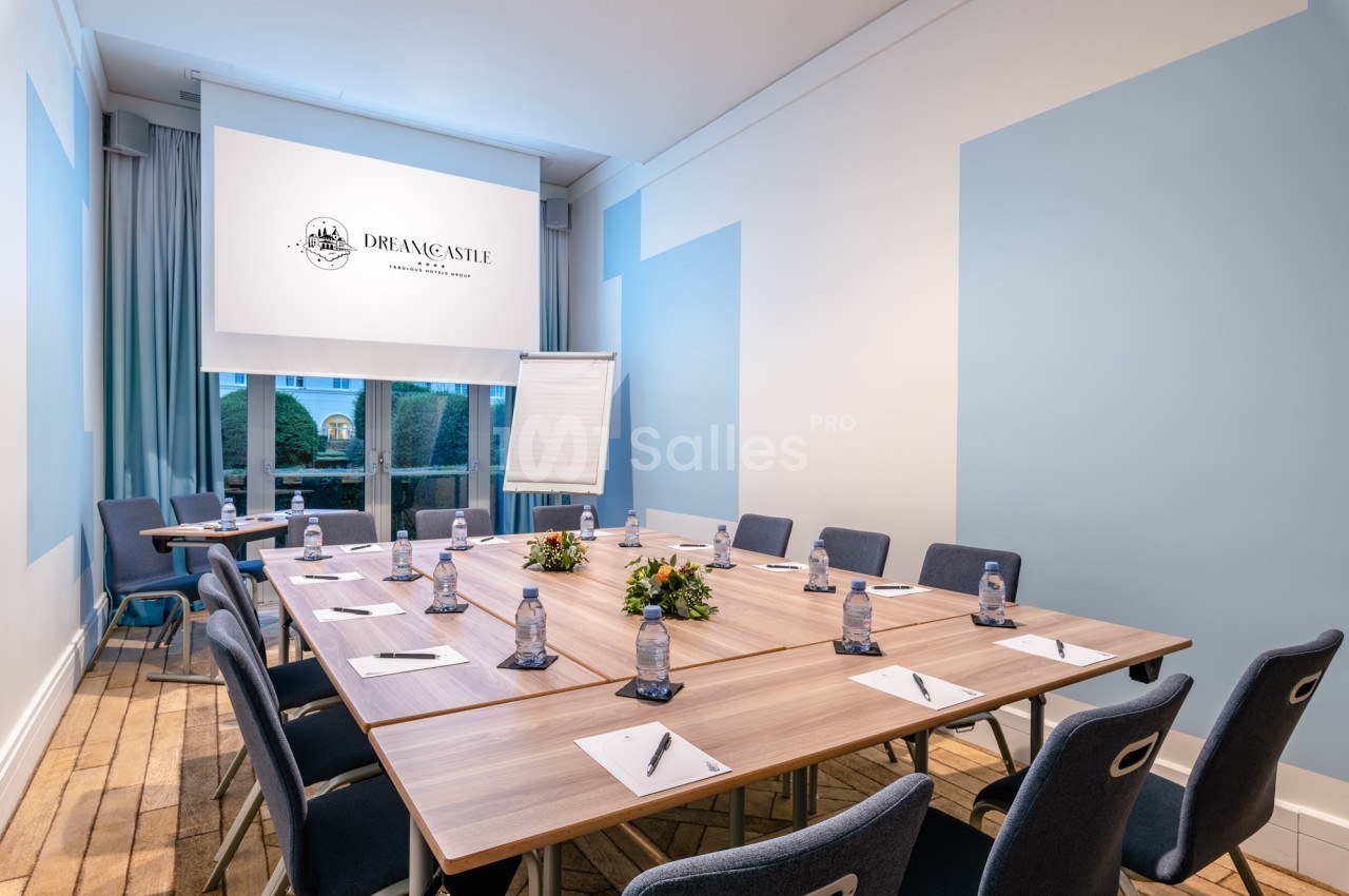 Salle de réunion lumineuse avec table en bois, chaises, bloc-notes, bouteilles d'eau et écran de projection.