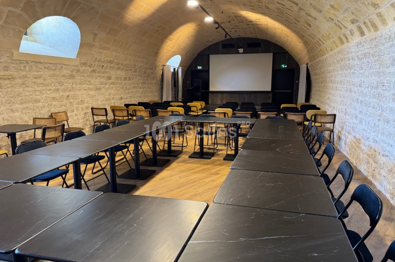 Salle de réunion voûtée avec murs en pierre, tables noires disposées en U et écran de projection au fond.
