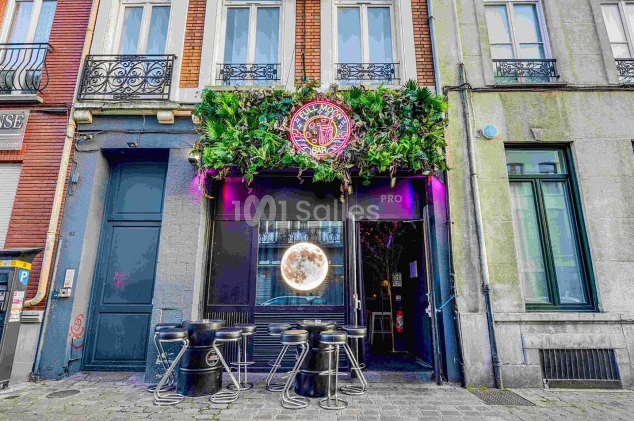Façade d'un bar avec une décoration végétale, des néons roses et des tabourets noirs sur le trottoir.