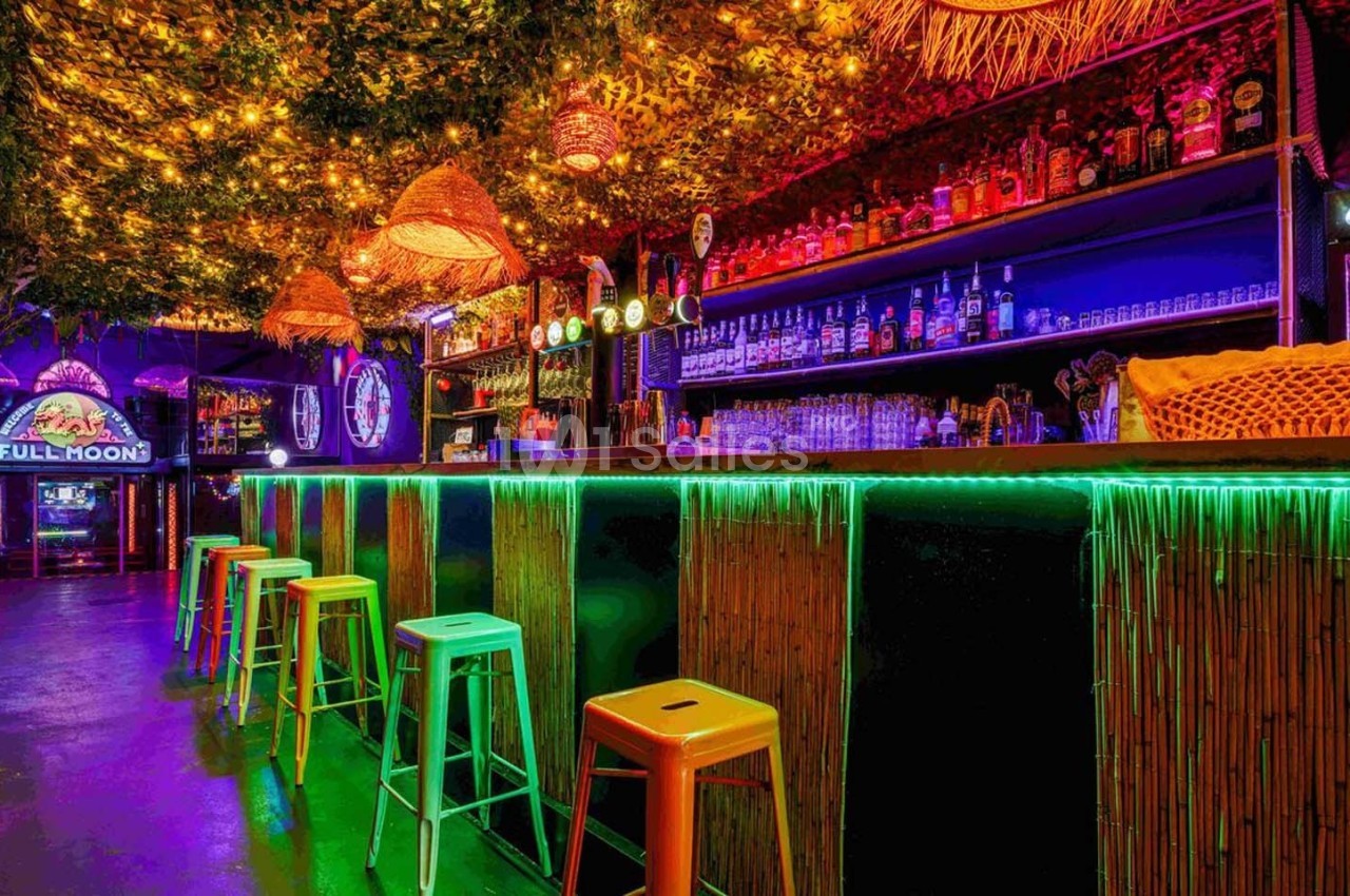 Bar coloré avec tabourets hauts, éclairage néon et plafond décoré de feuillage et lumières suspendues.