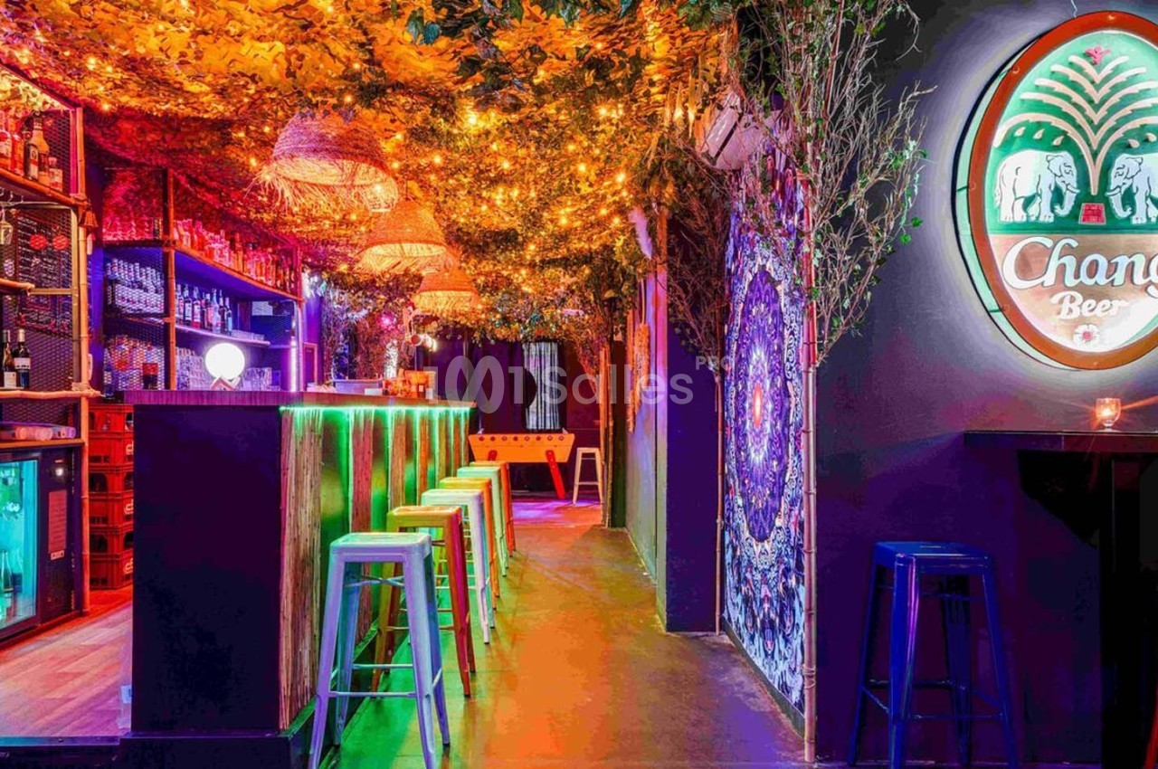 Bar coloré avec tabourets hauts, éclairage chaleureux, enseigne lumineuse et décor végétal au plafond.
