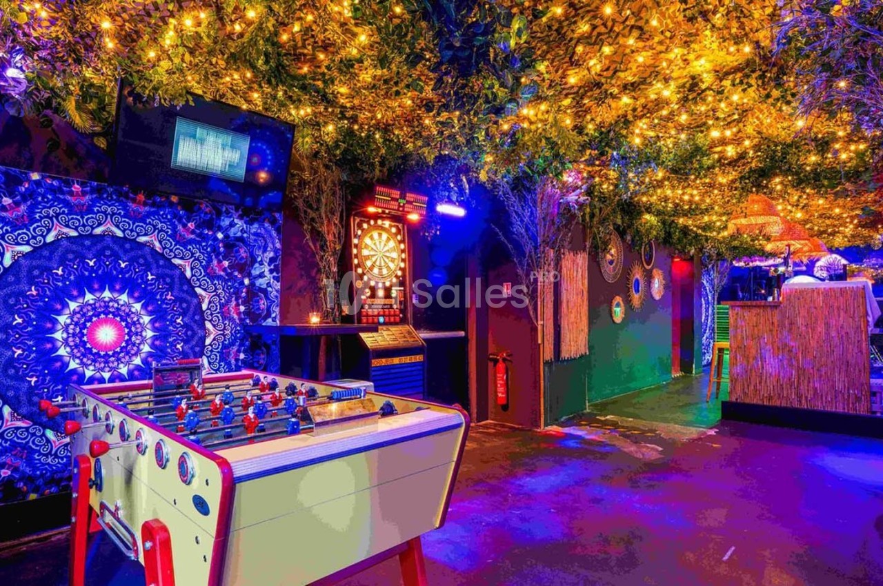 Salle de loisirs colorée avec baby-foot, cible de fléchettes, décorations lumineuses et végétation suspendue.