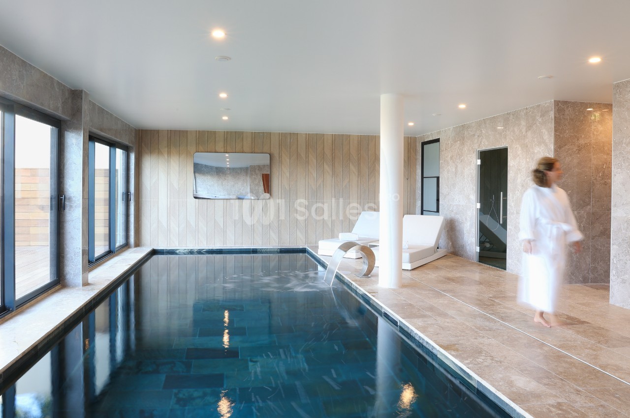 Piscine intérieure avec eau sombre, transats blancs, murs en bois et pierre, et une personne en peignoir flouement visible.