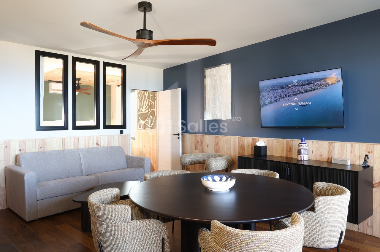 Salon moderne avec table ronde, chaises en rotin, canapé gris, mur bleu avec télévision et ventilateur au plafond.