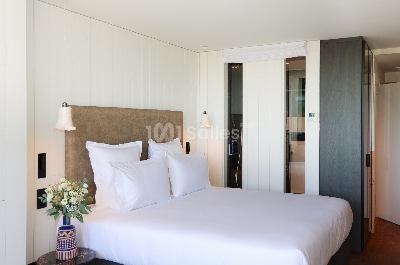 Lit double avec tête de lit rembourrée, draps blancs, lampes suspendues et vase de fleurs dans une chambre moderne.