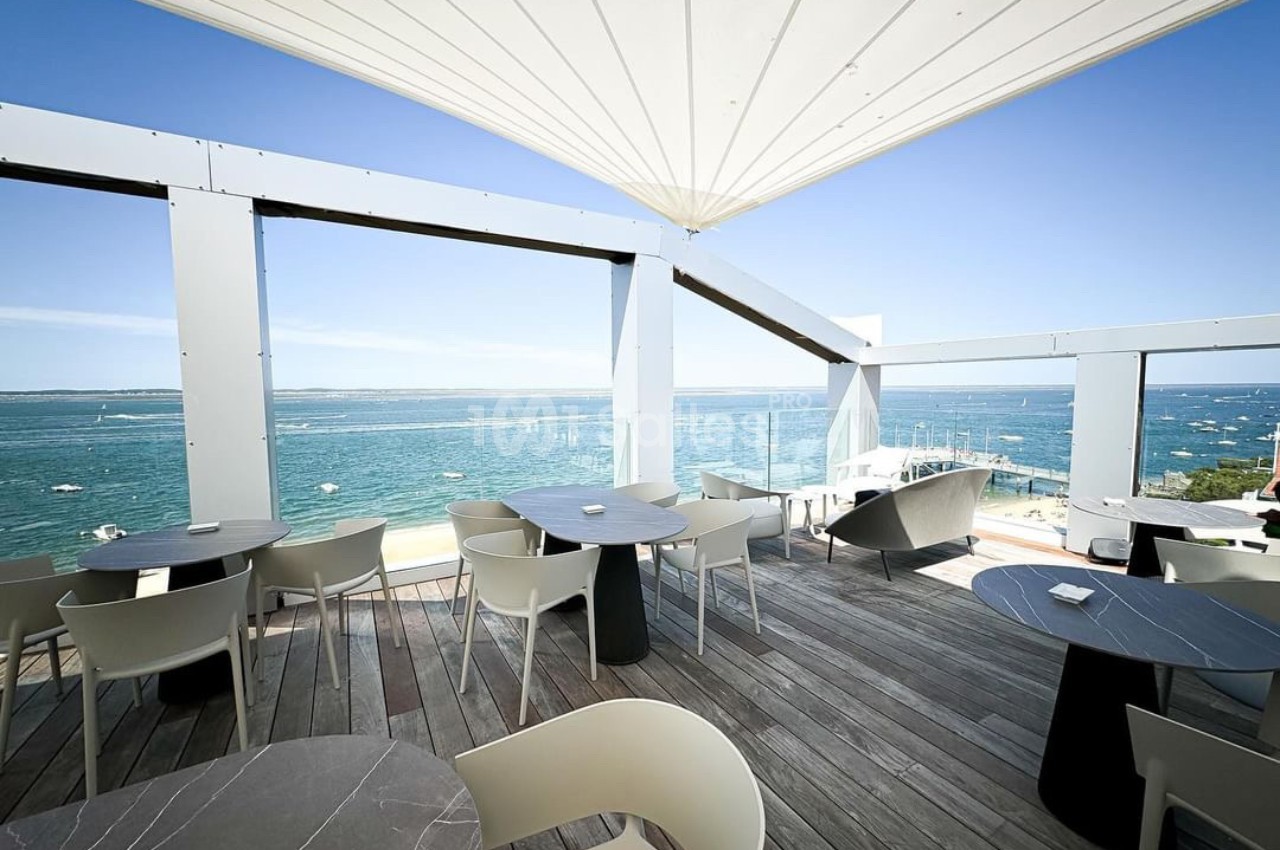 Terrasse en bois avec tables et chaises, offrant une vue dégagée sur la mer et un ciel bleu ensoleillé.