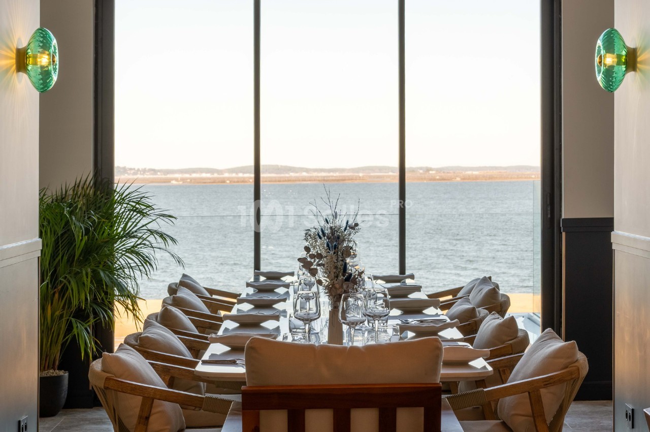 Salle à manger lumineuse avec une grande table dressée, vue sur un paysage de lac à travers de larges baies vitrées.