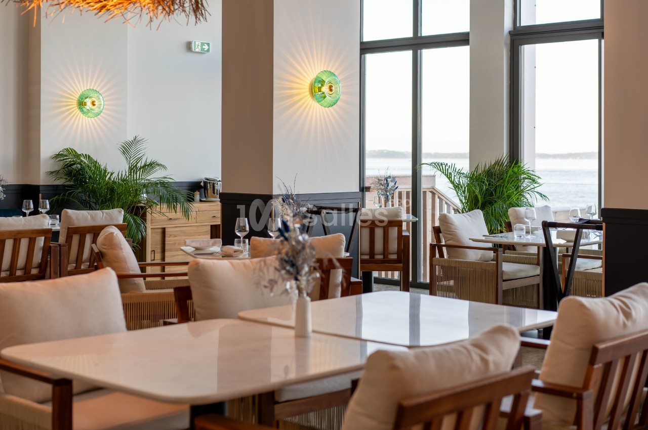 Salle de restaurant lumineuse avec tables en marbre, chaises en bois, plantes vertes et vue sur l'extérieur.