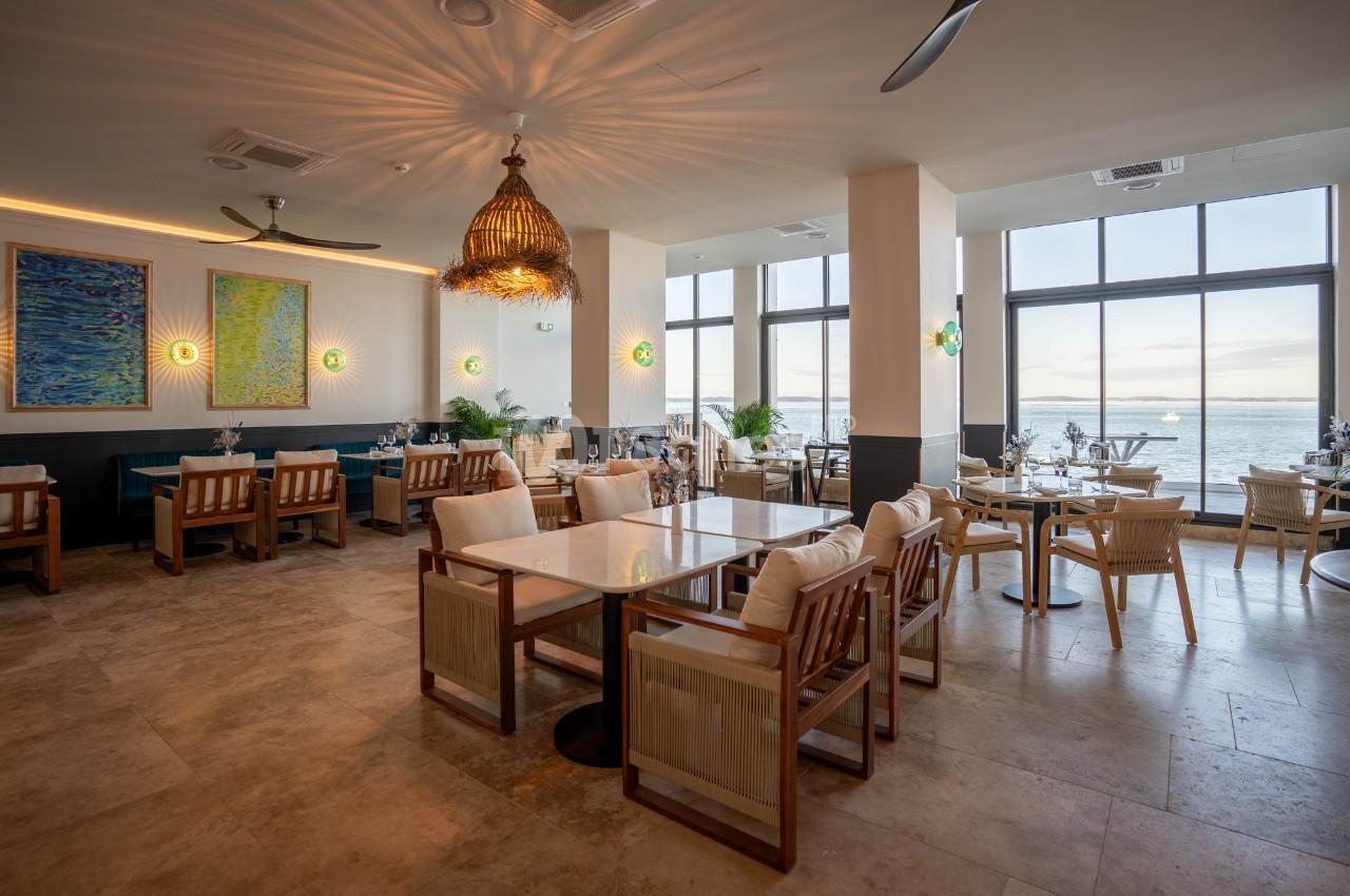 Salle de restaurant lumineuse avec grandes baies vitrées donnant sur la mer, tables et chaises en bois clair.