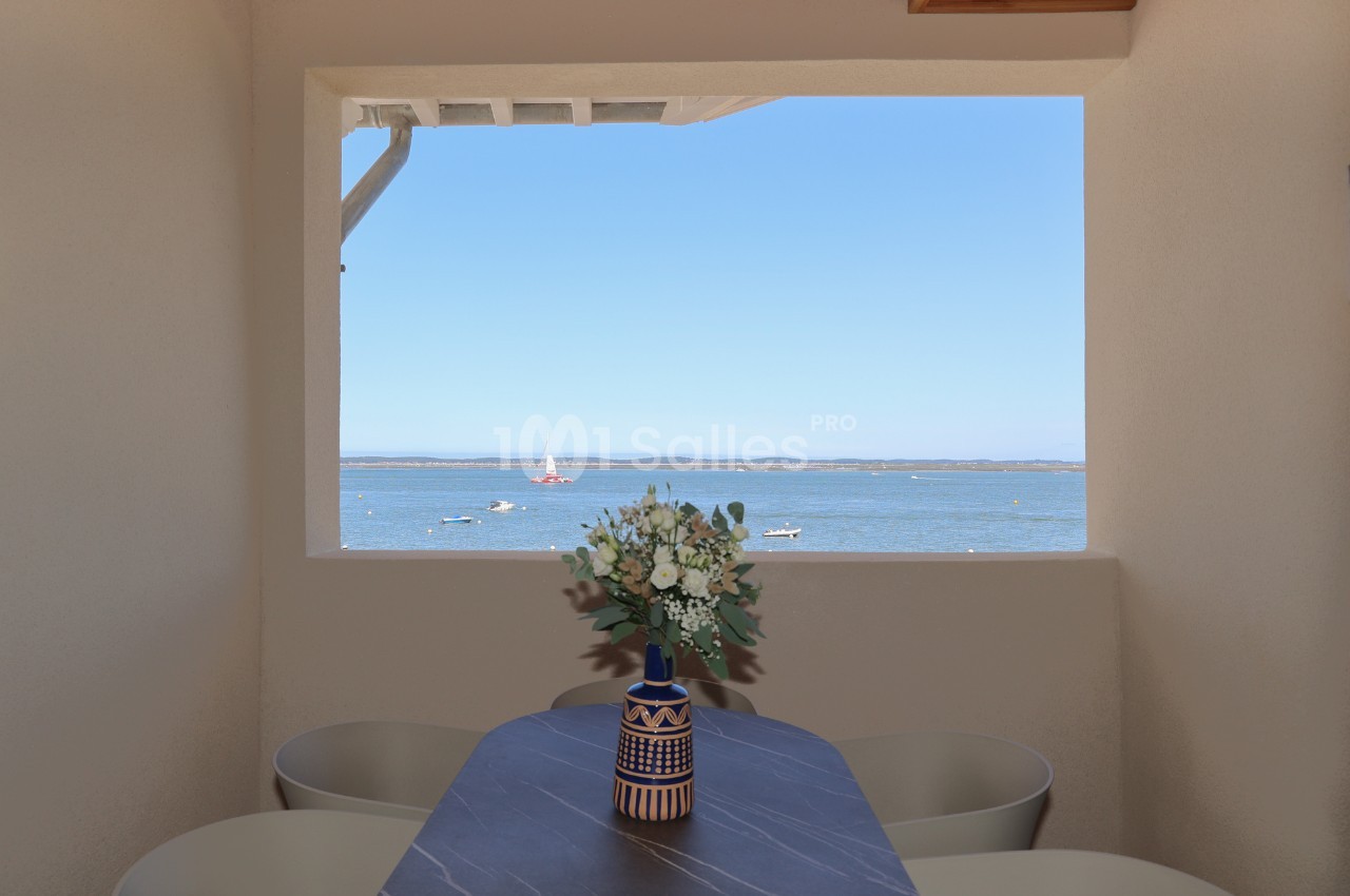 Vue sur la mer à travers une fenêtre encadrant un vase avec des fleurs sur une table en pierre sombre.