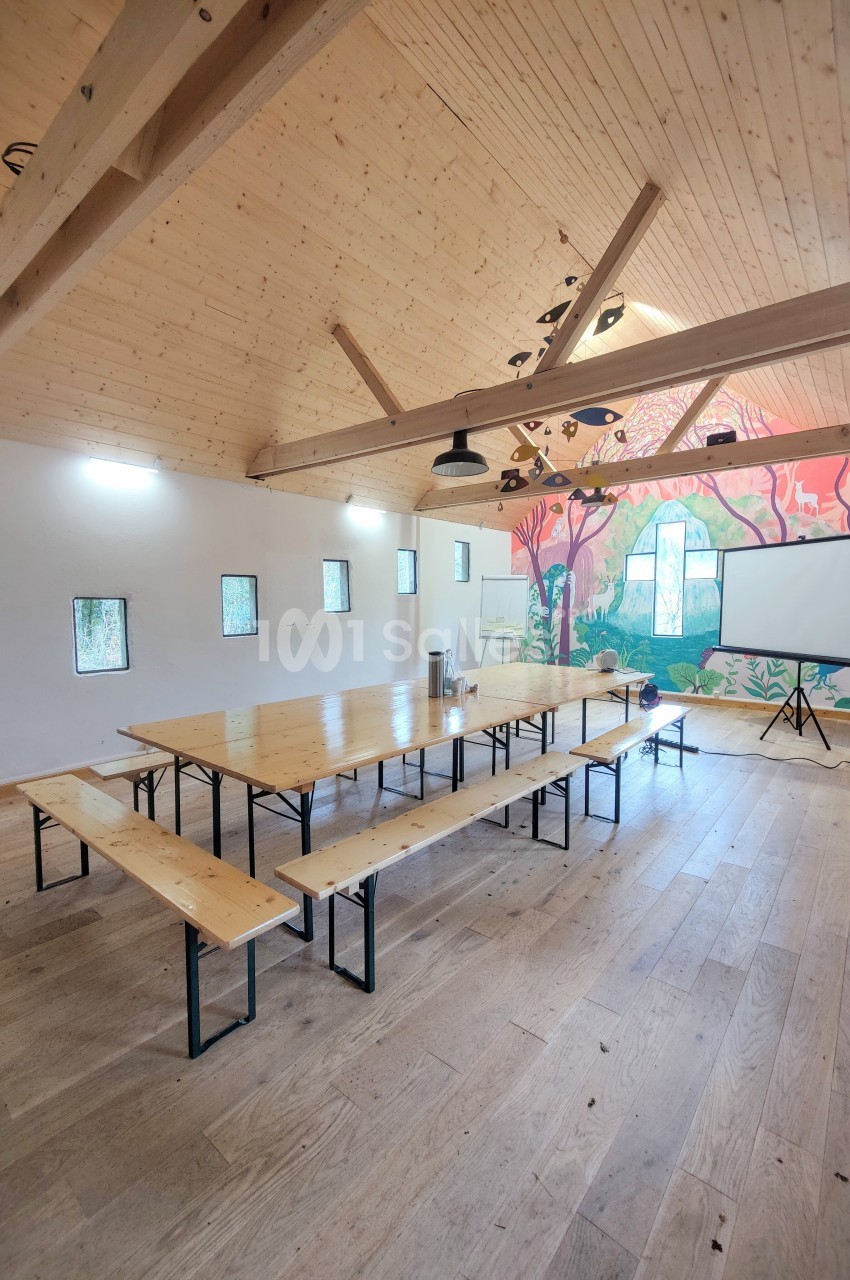 Salle lumineuse avec tables et bancs en bois, mur décoré d'une fresque colorée et écran de projection installé.