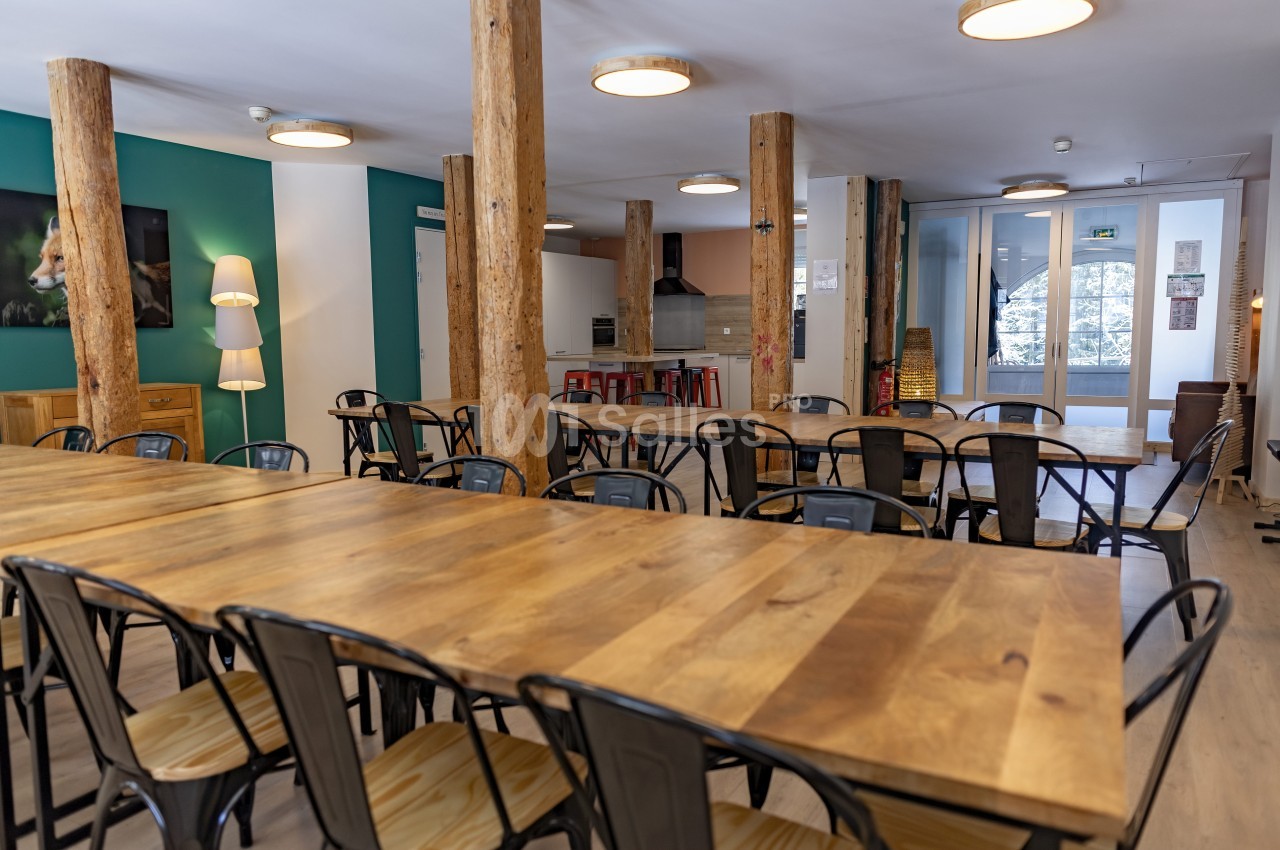 Salle lumineuse avec de grandes tables en bois, chaises métalliques, colonnes en bois et cuisine ouverte en arrière-plan.