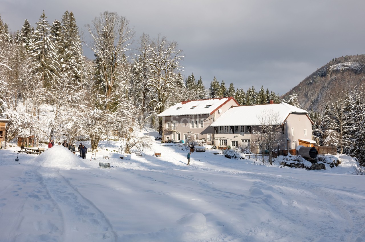 Paysage enneigé avec des bâtiments entourés d'arbres, des personnes profitant de la neige au premier plan.