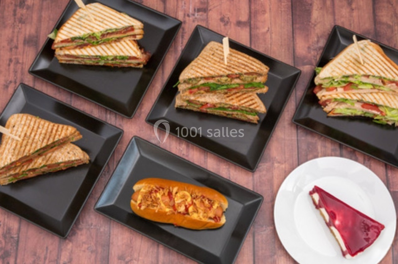 Assortiment de sandwichs grillés, un hot-dog et une part de gâteau sur des assiettes noires et blanches.