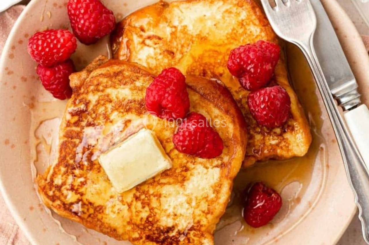 Deux tranches de pain perdu garnies de framboises, d'un morceau de beurre et de sirop, servies dans une assiette.
