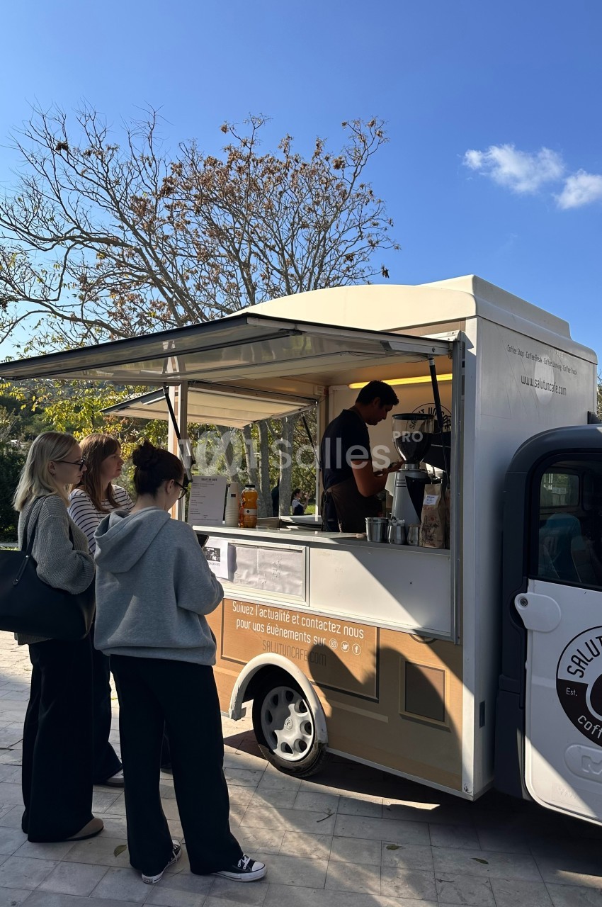 Un food truck sert des boissons chaudes à plusieurs personnes dans un espace extérieur ensoleillé.