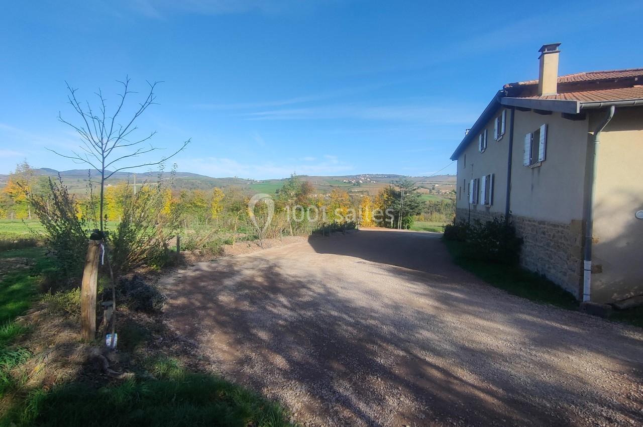 Chemin de gravier menant à une maison en pierre avec volets, entourée de végétation et d'un paysage rural.