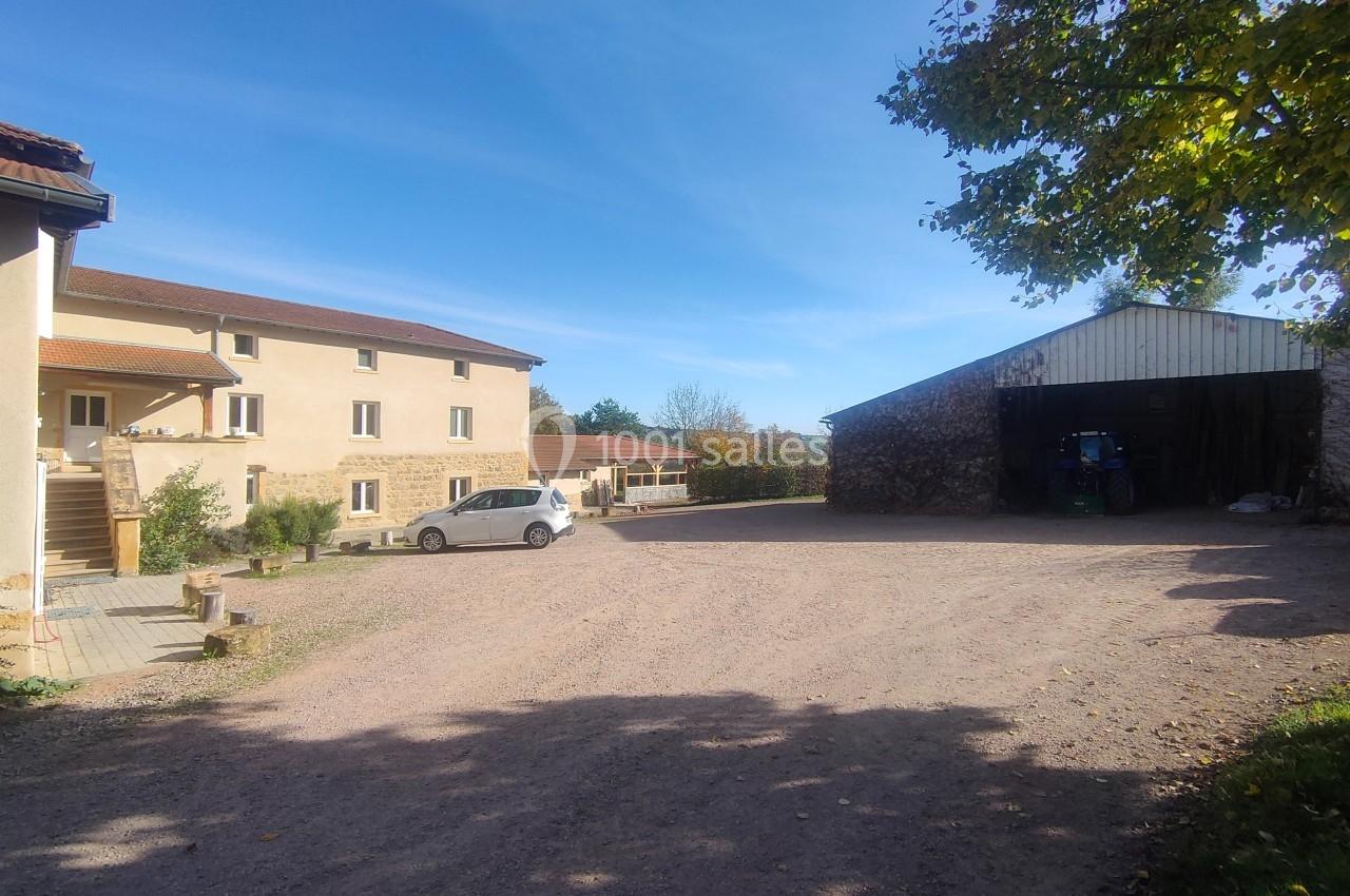 Cour d'une ferme avec bâtiments en pierre, un hangar ouvert, une voiture blanche et un ciel dégagé.
