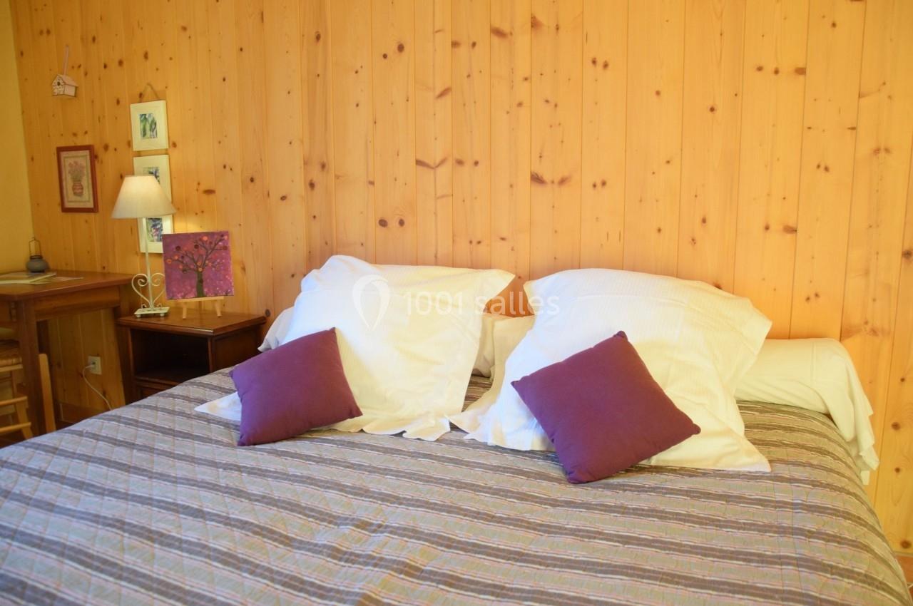 Lit double avec draps blancs, coussins violets et tête de lit en bois clair, dans une chambre au décor chaleureux.