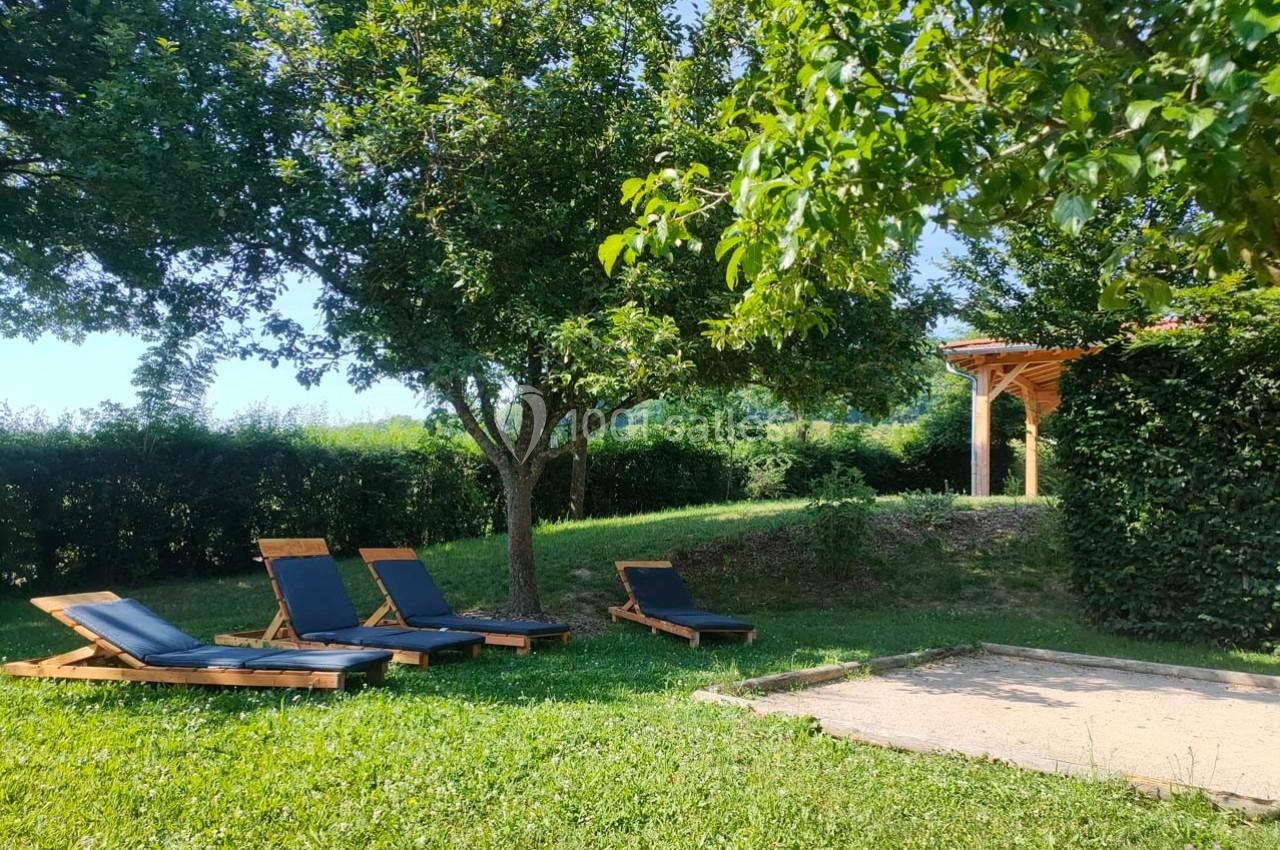 Quatre transats en bois sur une pelouse ombragée près d'un arbre et d'un terrain de pétanque, avec un abri en arrière-plan.